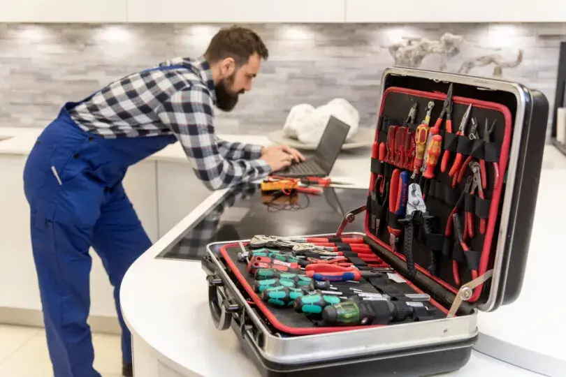 Handwerker steht in Küche und arbeitet am Laptop, Werkzeugkoffer mit Werkzeugen geöffnet.
